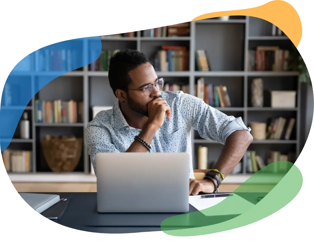 Man sitting at a desk with a laptop, thinking, in front of a bookshelf