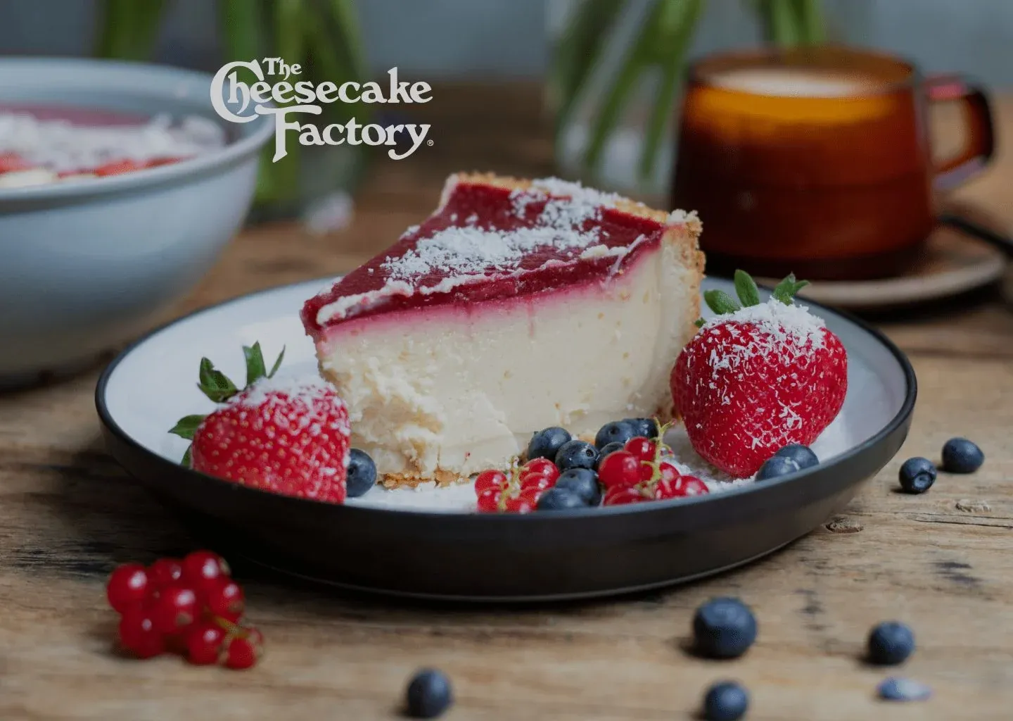 A slice of cheesecake with berry topping, strawberries, and blueberries on a dark plate, under The Cheesecake Factory logo
