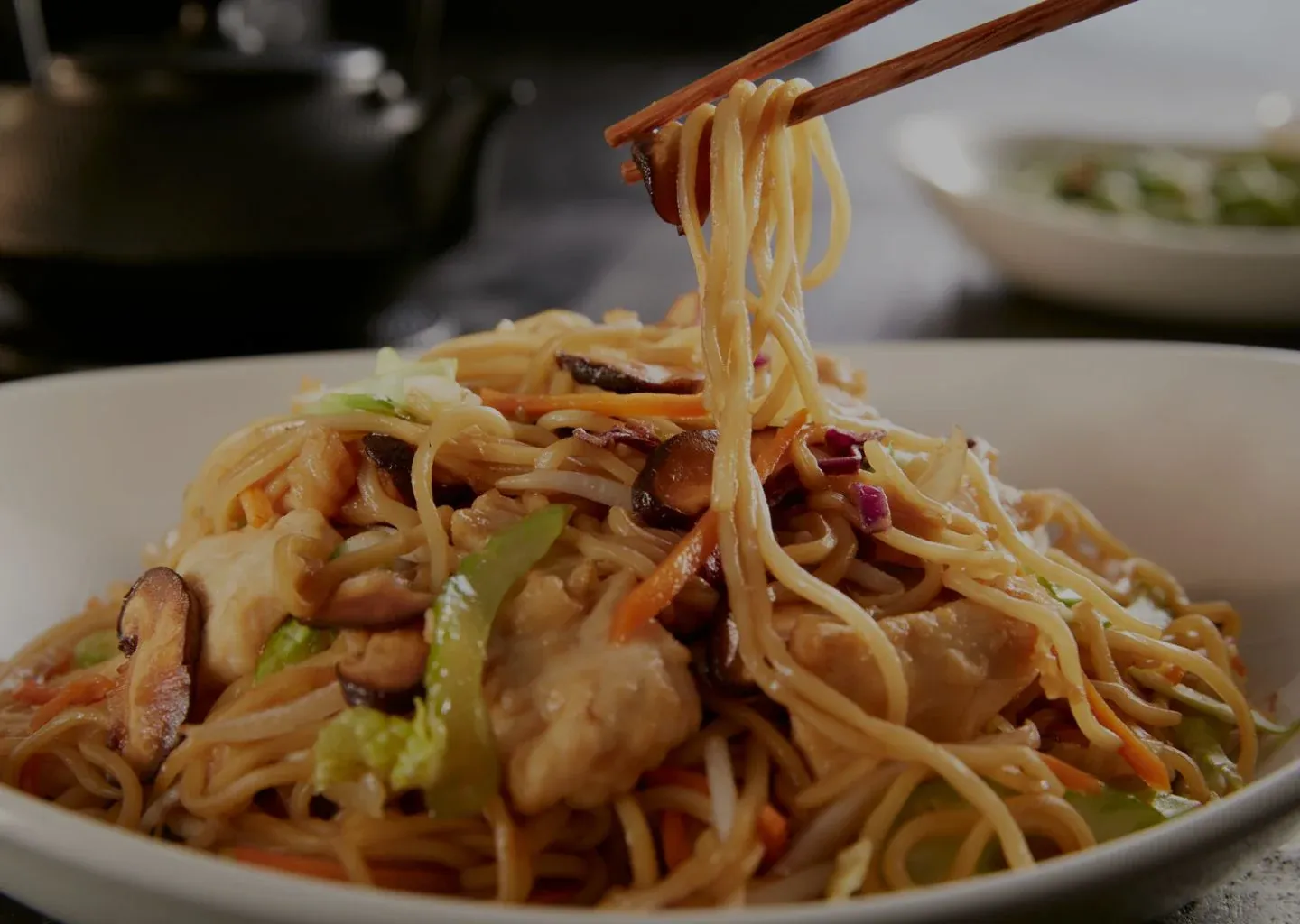 A plate of stir-fried noodles with chicken, mushrooms, and vegetables, lifted with chopsticks