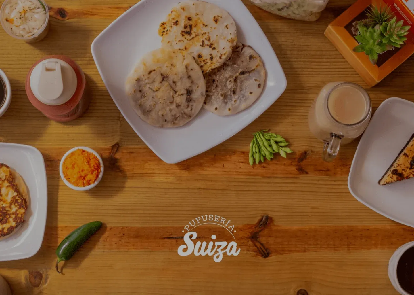Pupusas on white plates on a wooden table with salsa, curtido, and drinks, and the Pupusería Suiza logo