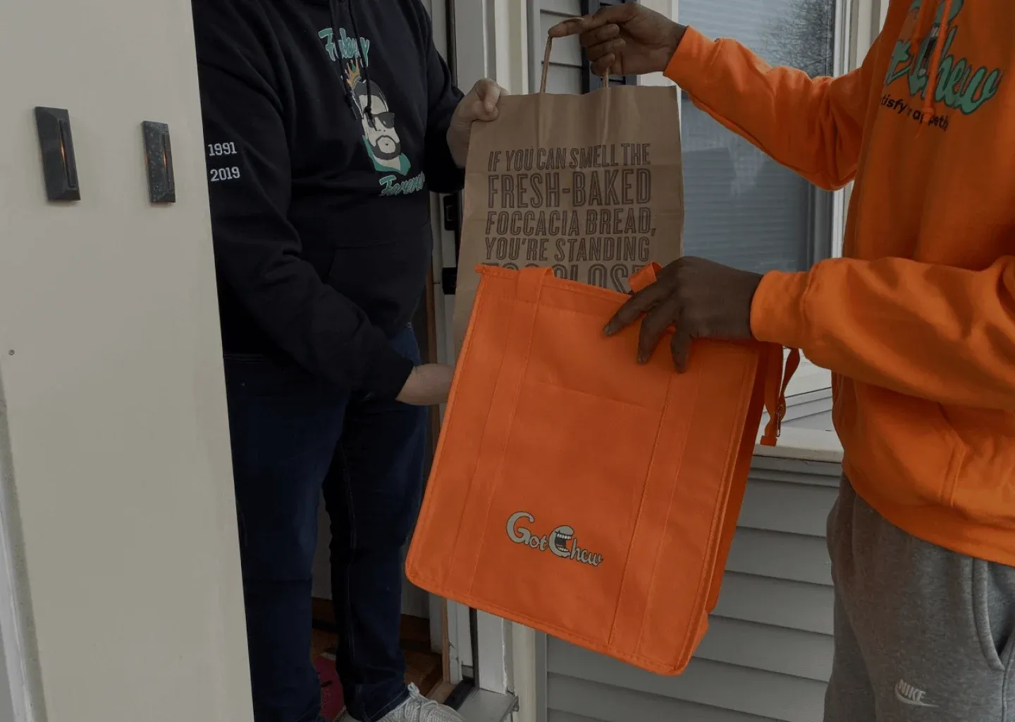 A GotChew courier in an orange hoodie handing a restaurant bag and an orange GotChew delivery bag to a customer at the door