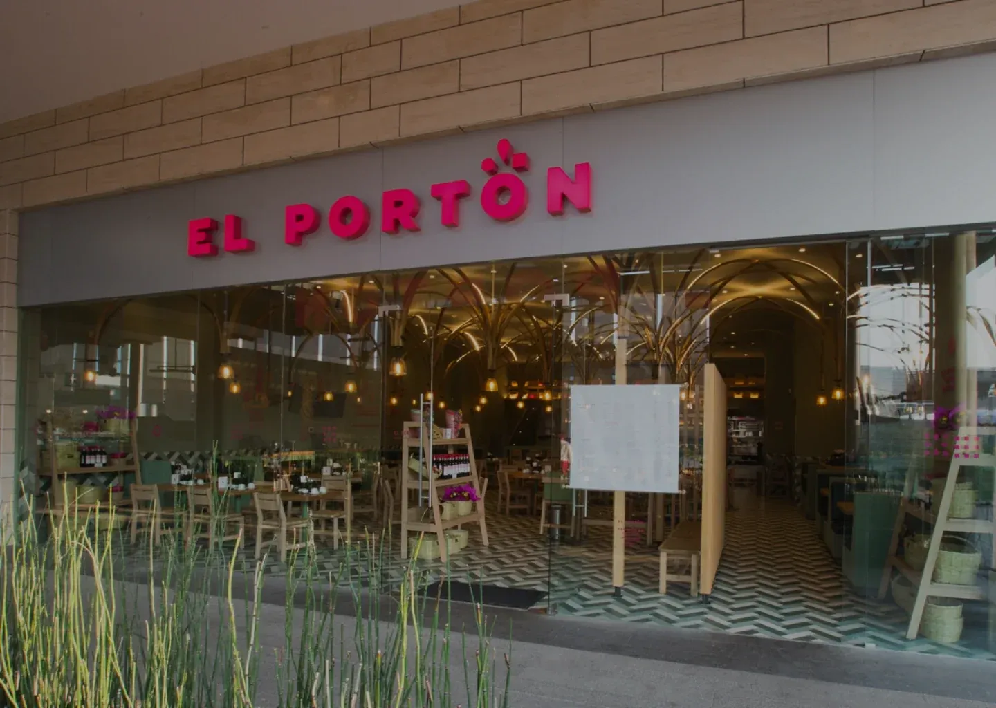 An El Portón restaurant with its pink sign above a glass front and plants along the entrance