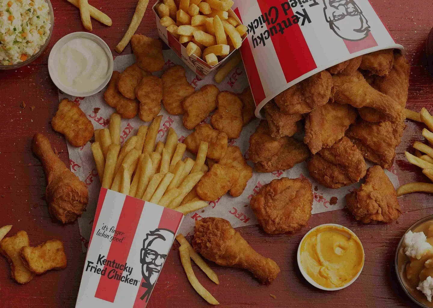 A KFC spread of fried chicken, nuggets, fries, and dips around a tipped-over Kentucky Fried Chicken bucket