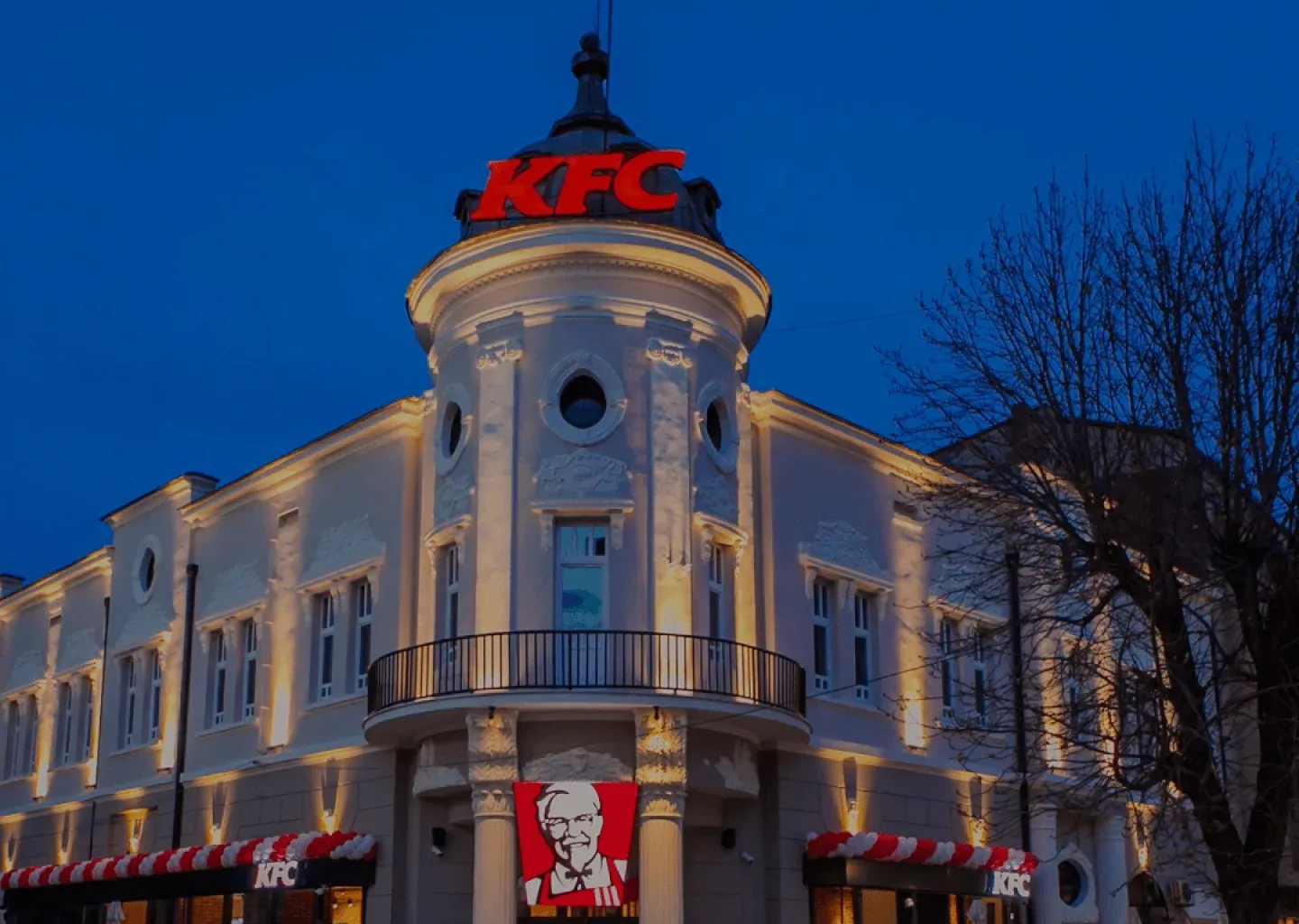 A KFC restaurant in a historic corner building at dusk, with a red KFC sign on its tower and the Colonel banner over the entrance