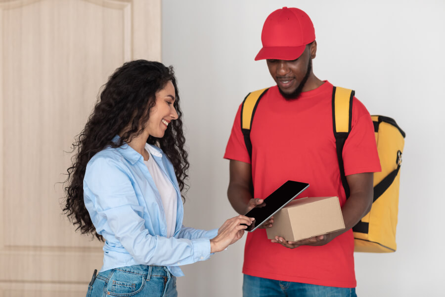 A delivery driver holds a package and a tablet while a customer signs on the tablet screen with her finger.