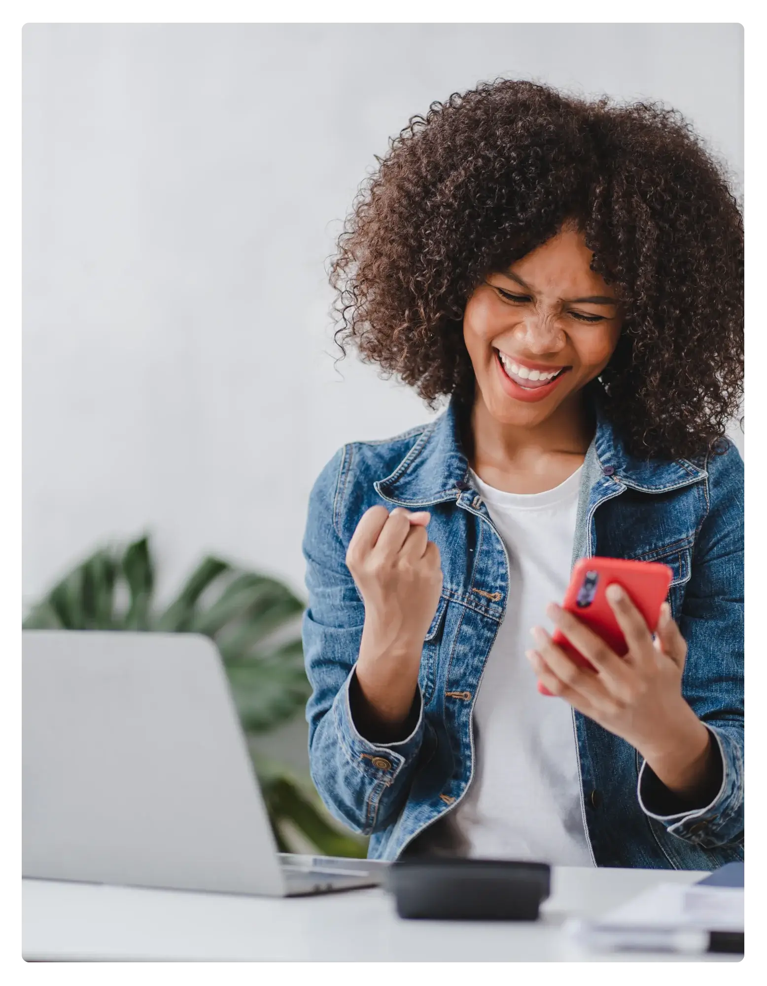 A woman in a denim jacket smiling and raising a fist while looking at her phone, with an open laptop and a plant on the desk.